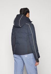 Veste en duvet marine avec capuche, dotée de passepoils blancs le long des coutures. Texture lisse avec une coupe ample et un petit logo de la marque sur l'ourlet.