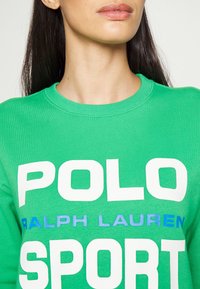 Sudadera verde de algodón con cuello redondo, con grandes letras blancas "POLO" y "SPORT" y un detalle azul en "RALPH LAUREN". Textura suave.