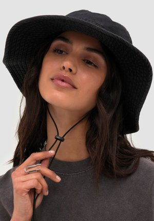 Femme aux longs cheveux foncés portant un chapeau noir à large bord et une chemise sombre ajuste la sangle sous le menton du chapeau avec sa main.