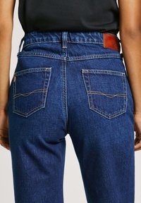Modré džínsy z denimu s vysokým pásom, elastickým pásom a dvoma zadnými vreckami s kontrastnými švami. Kožený značkový štítok na chrbte.