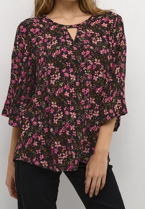 Kvinde iført en sort bluse med pink og lilla blomsterprint og en lille V-udskæring ved halsen, kombineret med sorte bukser.
