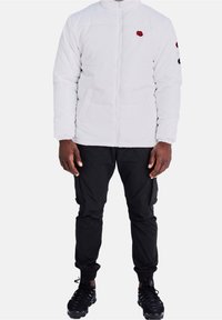 Man klädd i en vit jacka med dragkedja och röda lappar, svarta cargobyxor och svarta sportskor, stående mot en enfärgad vit bakgrund.