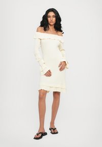Ribbed off-shoulder dress in off-white with long sleeves, ruffled edges, and a slightly flared hem paired with black flip-flops.