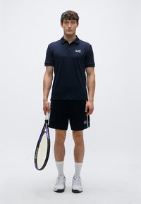 Tmavomodré polo tričko s logom, čierne šortky, biele ponožky a tenisová obuv. Drží fialovo-čiernu tenisovú raketu, stojí na jednoduchom pozadí.