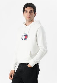 Weißer Hoodie mit einem Frontlogo, das rote und blaue Akzente aufweist. Hergestellt aus weichem Material, mit einer Kängurutasche und gerippten Bündchen.