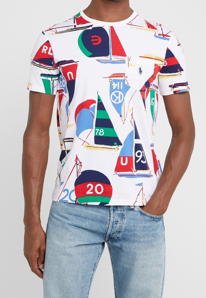 Vit t-shirt med ett färgglatt mönster av segelbåtar i blått, röd, grönt och svart. Rund halsringning och korta ärmar, tillverkad av mjuk bomull.