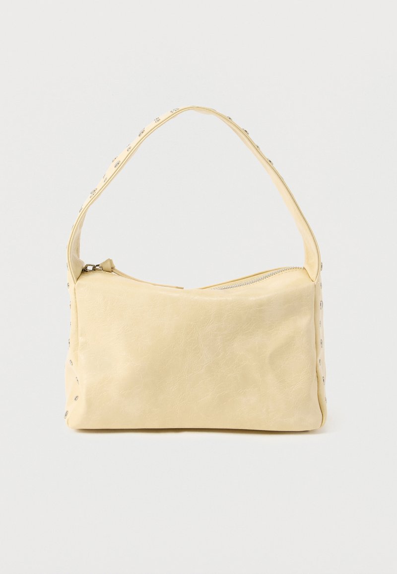 Μικρή μπεζ δερμάτινη τσάντα χειρός με έναν ιμάντα και ασημένιες τρουκ κατά μήκος των άκρων και του ιμάντα, τοποθετημένη πάνω σε απολύτως ουδέτερο φόντο.