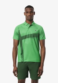 Polo verde de tela suave con un diseño de pinceladas en verde oscuro. Combinado con pantalones cortos verdes oscuros. Logo en el pecho.