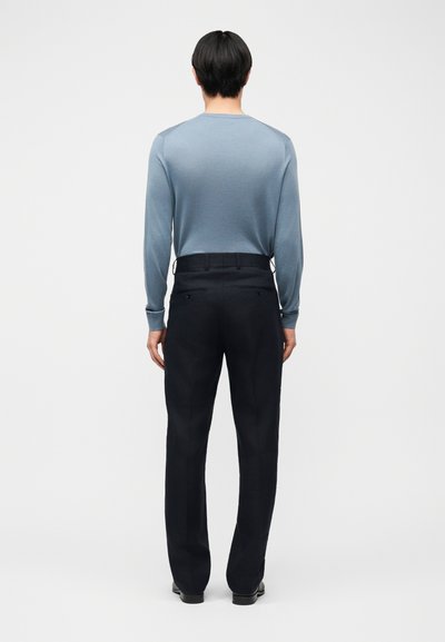 Άνδρας που στέκεται με πλάτη, φοράει ανοιχτό μπλε πουκάμισο με μακριά μανίκια και μαύρο παντελόνι φόρμας, σε απλό λευκό φόντο.