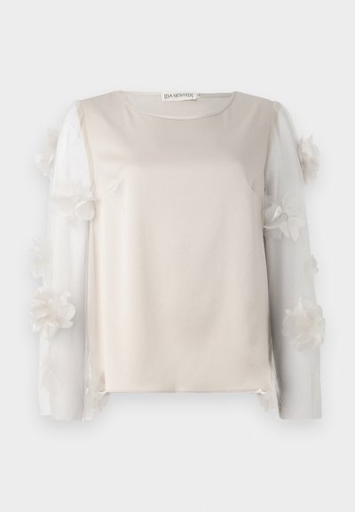Beige Oberteil mit einem glatten Stoffkörper und transparenten, mit floralen Akzenten versehenen Ärmeln, die an den Kanten geschichtete Blütendetails aufweisen.
