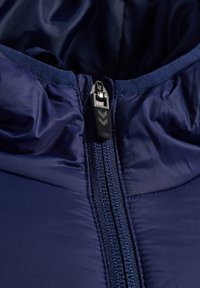 Marinblå vadderad jacka med en slät textur. Har en central dragkedja med en kontrasterande dragflik och sömmade detaljer runt kragen.