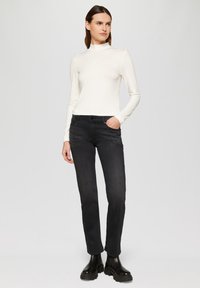 Jean noir en denim avec des embellissements sur les poches, associé à un haut ajusté à manches longues blanc. Le mannequin porte des bottes noires épaisses.