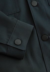 Mørkegrøn jakke med tekstureret stof, der har en knappet manchet og sorte metalknapper. Lodrette striber er synlige i materialet.