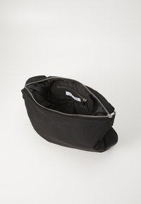 Bolsa de nailon negra con un zipper plateado. Diseño suave y flexible con un bolsillo interior y marca. Textura suave y apariencia minimalista.