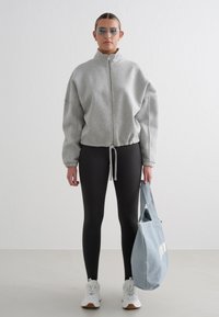 Lysegrå hettegenser med glidelås og høy krage, kombinert med svarte tights, hvite joggesko og en blå lerretsveske med hvit tekst.