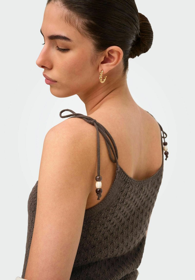 Jeune femme aux cheveux en chignon porte un haut sans manches texturé foncé avec des bretelles nouées aux épaules et des boucles d’oreilles créoles dorées, regardant de profil les yeux fermés.
