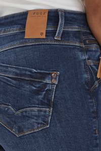 Pantalones vaqueros de mezclilla en azul oscuro con una etiqueta de cuero de la marca. Las características incluyen costuras en contraste, bolsillos traseros con remaches decorativos.