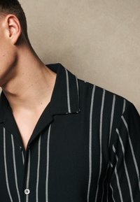 Camisa negra de cuello con rayas verticales blancas, textura acanalada y un detalle de botón en el cuello, que muestra un diseño moderno.