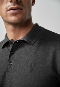 Mørkegrå polo-shirt lavet af tekstureret stof, med en klassisk krave, tre knapper og et diskret logo på venstre side.