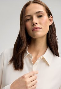 Blusa crema con colletto a punta, chiusura con bottoni frontale e un abbellimento decorativo vicino al colletto. Tessuto dalla consistenza liscia e design aderente.