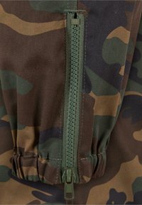 Zöld és barna árnyalatú, terepmintás anyag, kiemelkedő zöld cipzárral, alul összegyűrt derékkal.