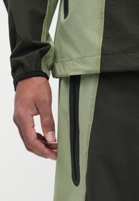 Heren sportkleding met een twee-kleurig ontwerp in lichtgroene en donkere olijfkleuren, elastische manchetten en zijzakken met rits.