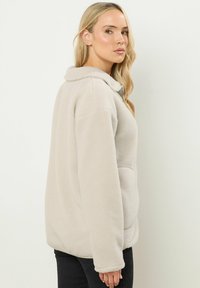 Giacca in pile color beige chiaro con colletto, maniche lunghe e tasca frontale. Texture morbida con vestibilità rilassata, mostrata da un angolo laterale.