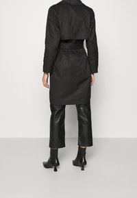 Cappotto lungo nero con design accorciato, vita con cintura, maniche lunghe e una texture liscia. Abbinato a pantaloni di pelle e stivali con tacco.