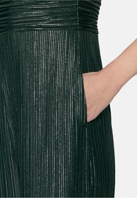 Robe verte à rayures métalliques verticales, avec un corsage froncé et une poche latérale. Le tissu a une finition lisse et texturée.