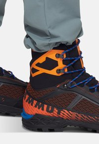 Bottines de randonnée noires et orange avec une tige texturée, des lacets bleus et une semelle renforcée avec des motifs distinctifs et une marque.