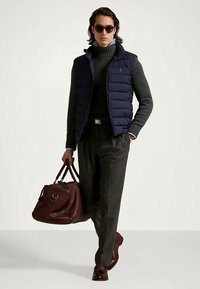 Άντρας με σκούρα γυαλιά ηλίου, ναυτική επενδυμένη γιλέκα, γκρι ψηλόλαιμο πουλόβερ, γκρι παντελόνι, καφέ παπούτσια, κρατώντας μια καφέ δερμάτινη τσάντα.