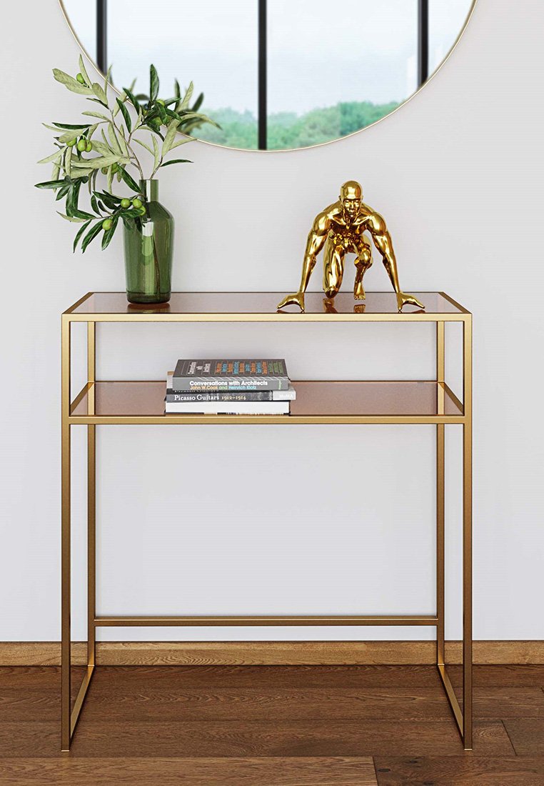 Konsolentisch aus goldenem Metall mit zwei transparenten Glaselementen; zeigt eine skulptierte goldene Figur und eine grüne Vase, die mit Laub gefüllt ist.