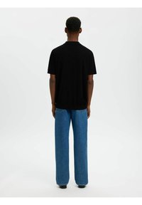 Homme debout, de dos, portant une chemise noire à manches courtes et un jean large bleu, sur un fond blanc uni.