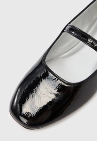 Bailarina de charol negro con punta cuadrada y una sola correa sobre el empeine. Textura suave y diseño elegante. Forro plateado visible.