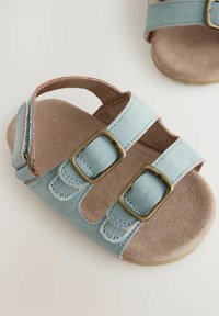 Sandal in ecopelle blu chiaro con due cinturini regolabili, fibbie in bronzo e una soletta beige morbida. Suola piatta con dettagli testurizzati.