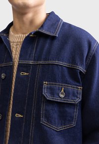 Dunkelblaue Jeansjacke mit Knopfleiste, gelben Nähten und einer Brusttasche. Sie verfügt über einen klassischen Kragen und einen strukturierten Strickpullover darunter.