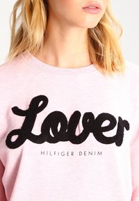 Roze sweatshirt met een groot zwart geborduurd "Lover"-tekst en daaronder een kleinere "HILFIGER DENIM". Zachte stof met een ronde hals ontwerp.