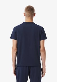 Hombre de pie, de espaldas, vistiendo una camiseta de manga corta azul marino lisa y pantalones azules marinos a juego, sobre un fondo blanco.