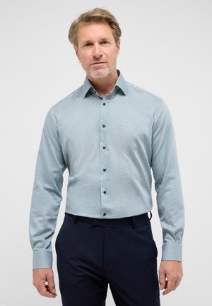 Hellblaues Hemd mit Knöpfen und geometrischem Muster, mit abgerundetem Kragen und dunklen Knöpfen, kombiniert mit marineblauen Hosen.