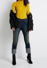 Gelbes Rollkragenoberteil, schwarze Jacke, dunkelblaue Jeans mit leichter Ausbleichung und hochgekrempelten Säumen, schwarze Stiefeletten mit Reißverschlüssen und hohen Absätzen.