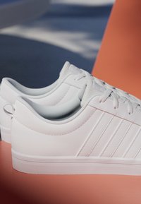 Par de zapatillas bajas blancas con tres franjas laterales, cordones blancos y suelas de goma texturizadas sobre una superficie naranja.