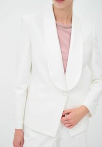 Blazer bianco su misura con collo sciallato, chiusura con un singolo bottone e una tasca frontale. Indossato sopra una maglietta rosa chiaro. Tessuto liscio.