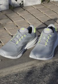 Chaussures de sport grises avec une tige en maille texturée, des accents jaune fluo et trois bandes sur les côtés. Semelles en caoutchouc et design à lacets.