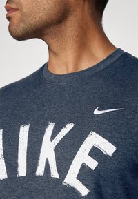 Námořnická modrá tričko s kulatým výstřihem vyrobené z měkké látky, které má velké bílé písmeno "NIKE" a malé bílé logo Nike swoosh na hrudi.