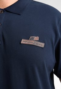 Mörkblå pikétröja i textil med struktur, med en krage med knappar och en broderad "International Speedway"-logga med detaljer.