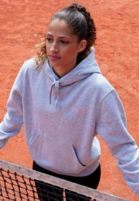 Felpa grigia con cappuccio e tasca frontale, indossata sopra leggings neri, posizionata su uno sfondo di un campo da tennis in argilla rossa.