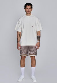 T-shirt en coton blanc avec un petit logo rouge, associé à un short en satin marron clair avec une bordure dorée, des chaussettes blanches et des baskets blanches.