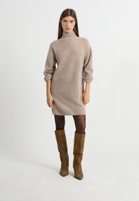 Vestido de punto beige con cuello alto y mangas tres cuartos, con bordes acanalados. Combinado con botas de ante marrón hasta la rodilla y medias negras.