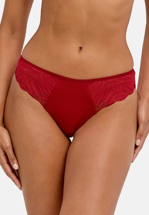 Kvinde iført rød blonde- og stof-bikini, der viser midten af kroppen og øverste lår mod en lys baggrund.