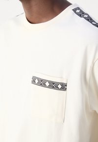 Cream t-shirt with a chest pocket featuring a black geometric pattern. Soft fabric, standard crew neck, and contrasting shoulder details.